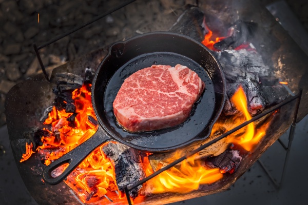 A large steak cooking in a cast iron pan over a campfire