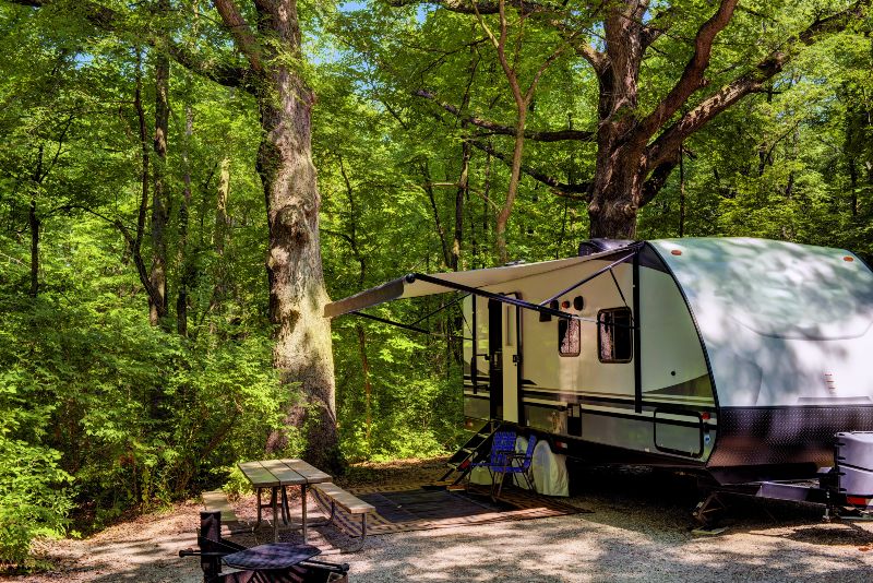 An RV with an extended awning parked next to a bench in the woods